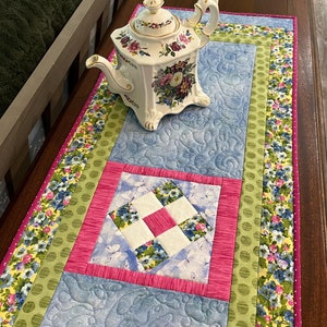 May include: A quilted table runner featuring a floral design in shades of pink, green, blue and yellow. The runner has a square patchwork design in the centre and a scalloped edge. A white teapot with floral decorations is sitting on the runner.