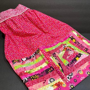 Pode incluir: Uma saia rosa com bolinhas brancas e uma bainha de patchwork colorida. A bainha apresenta padrões florais e geométricos em tons de rosa, verde e marrom.