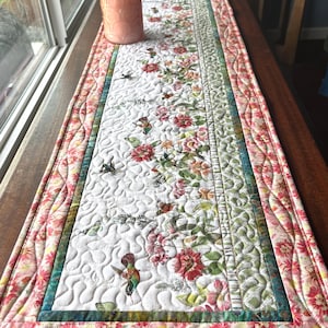 May include: A rectangular quilted table runner featuring a white central panel with a printed pattern of red flowers, green leaves, and hummingbirds. The runner is bordered by a floral fabric in shades of pink and yellow with wavy stitching.