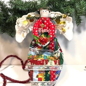 May include: A handmade Christmas angel tree topper. The angel has a fabric body with a patchwork design featuring holiday motifs. It wears a red top with white polka dots and holds a bouquet. White wings and a halo complete the festive look.