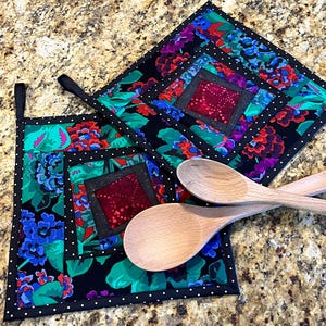May include: Two square quilted pot holders with a floral pattern in shades of blue, green, red, and purple on a black background. The pot holders have a black border with white polka dots. Two wooden spoons are resting on the pot holders.