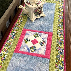 May include: A floral teapot sits on a quilted table runner. The runner features a light blue center with a pink and white square design. The edges are bordered with green polka dots and floral patterns. The teapot is white with floral designs.