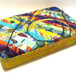 May include: A rectangular, fabric-covered box with a vibrant, patchwork design. The lid features a colorful array of fabric strips in various colors, with decorative stitching. The box has a yellow base and a button closure. The word "Inspire" is visible.