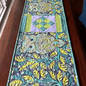 以下が含まれることがあります： 花柄とフクロウのデザインのキルトテーブルランナー。ランナーは、黄色とティール色の葉、ティール色のフクロウ、ラベンダーと緑のダイヤモンド模様の灰色の背景が特徴です。端はティール色の水玉模様の生地でトリミングされています。