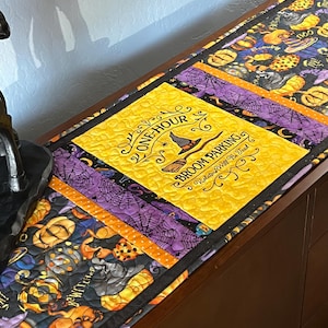 May include: A quilted table runner featuring a yellow central panel with the text ONE-HOUR BROOM PARKING Violators Will Be Toad, surrounded by sections of black fabric printed with orange pumpkins, purple spiderwebs, and witch hats.