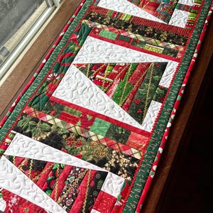 May include: A rectangular quilted table runner featuring a repeating pattern of stylized Christmas trees. The trees are constructed from various fabric strips in red, green, and black with holiday prints, set against a white stippled background.