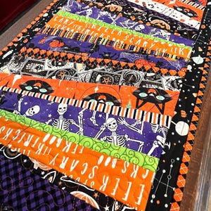 May include: A rectangular quilted table runner featuring a patchwork design of Halloween-themed fabrics in orange, purple, green, and black. The fabric patterns include skeletons, black cats, pumpkins, and text reading EEEK SCARY TRICKS.