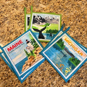 Peut inclure: Trois maniques carrées en tissu avec des illustrations imprimées de l'Alaska avec une queue de baleine, du parc national d'Acadia dans le Maine avec un phare, et des Grands Lacs du Michigan. Chaque pièce a une bordure en tissu à pois verts ou bleus.