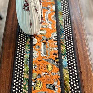 May include: A black and white polka dot table runner with a black border and a colorful Halloween themed center panel. The center panel features a variety of Halloween imagery, including pumpkins, bats, spiders, and ghosts. The text "Trick or Treat" and "Halloween" are also visible on the panel.
