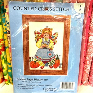 May include: A Counted Cross Stitch kit titled Kitchen Angel Picture 5117 by Candamar Designs. The packaging features a cross-stitched angel with orange hair, yellow sleeves, and a blue apron holding a pie, surrounded by pumpkins and corn, in a 5" x 7" frame.