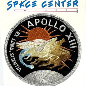 Op de afbeelding: Een ronde geborduurde patch met het Apollo XIII missie-embleem. Het ontwerp toont drie paarden die voor een zon rennen, tegen een zwarte achtergrond met de tekst "APOLLO XIII" en "EX LUNA, SCIENTIA". De patch is bevestigd op een witte kaart met het opschrift "SPACE CENTER HOUSTON".