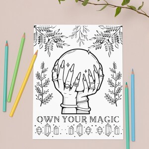 May include: Black and white illustration of hands holding a crystal ball, with the text "OWN YOUR MAGIC". The image is surrounded by botanical elements and decorative crystals. Colored pencils in blue, green, and yellow are placed around the image.