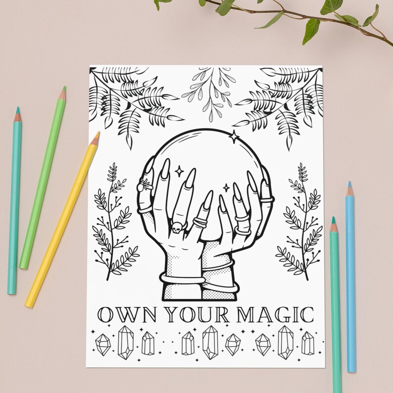 May include: Black and white illustration of hands holding a crystal ball, with the text "OWN YOUR MAGIC". The image is surrounded by botanical elements and decorative crystals. Colored pencils in blue, green, and yellow are placed around the image.