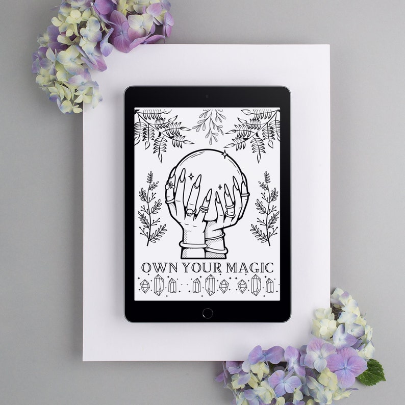 May include: A black tablet displays a black and white illustration of hands holding a crystal ball, surrounded by botanical elements. The text "OWN YOUR MAGIC" is below the illustration. The tablet is set on a white surface, with purple flowers in the corners.
