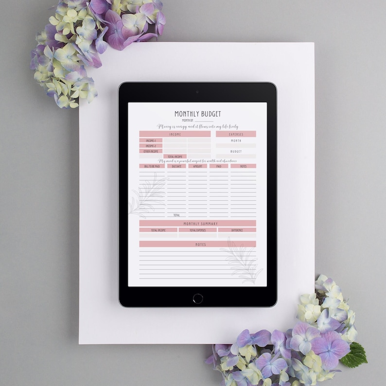 K&ouml;nnte beinhalten: Ein digitales Tablet zeigt einen druckbaren monatlichen Haushaltsplaner mit Abschnitten f&uuml;r Einnahmen, Ausgaben und eine monatliche Zusammenfassung. Der Planer hat ein rosa-wei&szlig;es Farbschema mit floralen Akzenten.