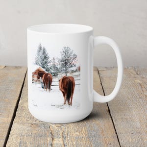 May include: White ceramic mug featuring a winter scene with two brown horses in a snowy field near a wooden structure. The horses are facing away from the viewer. Snow-covered trees and a fence are in the background. The mug has a curved handle.