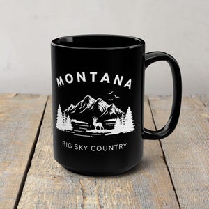 May include: Black ceramic mug with a white graphic of a mountain range, trees, and a deer. The word "MONTANA" is above the graphic, and "BIG SKY COUNTRY" is below it. The mug has a large handle.