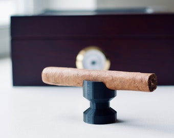 May include: A brown cigar rests on a black, geometric cigar stand. A dark wooden humidor with a gold-colored hygrometer is in the background. The cigar is a light brown color.