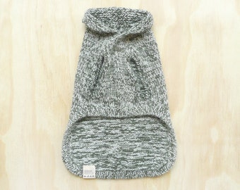 May include: A sleeveless pet sweater featuring a marled knit pattern in shades of sage green and white. The garment has a folded turtleneck collar, two arm openings, and a curved hemline. A small white care label is attached to the bottom left corner.