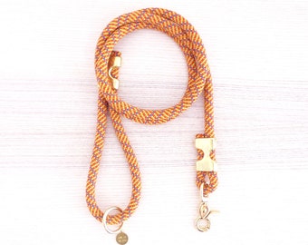 Peut inclure: Laisse pour chien en corde orange et bleue à motif avec une boucle en métal doré et un anneau en métal doré. La laisse est sur une surface en bois blanc.