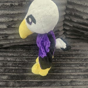 May include: A plush eagle toy featuring a white head, a yellow beak, and purple and black wings. The stuffed animal has a small white and black tail and yellow feet. The eyes are embroidered with purple and black thread on the white plush head.