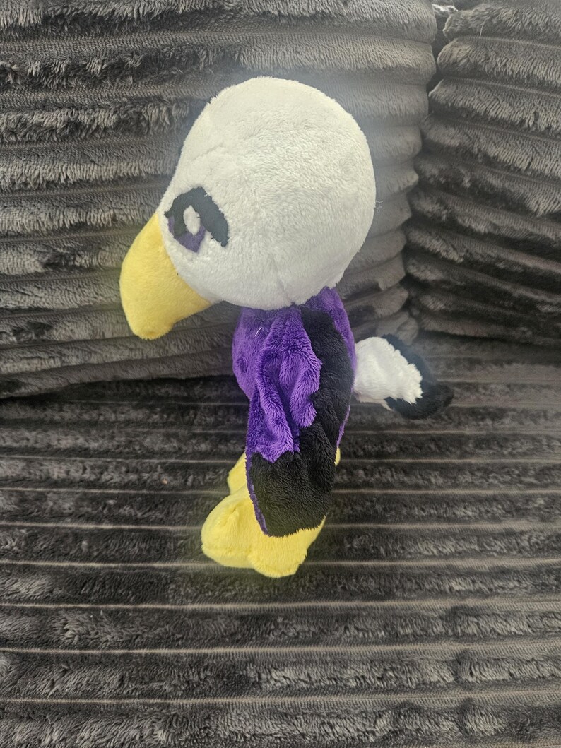 May include: A plush eagle toy featuring a white head, a yellow beak, and purple and black wings. The stuffed animal has a small white and black tail and yellow feet. The eyes are embroidered with purple and black thread on the white plush head.