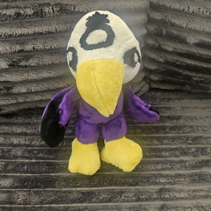 May include: A plush toy shaped like a bird with a white head, a yellow beak, and yellow feet. The body is purple with black wing tips. A black circular symbol is embroidered on the forehead. The eyes are dark with purple accents on the sides.