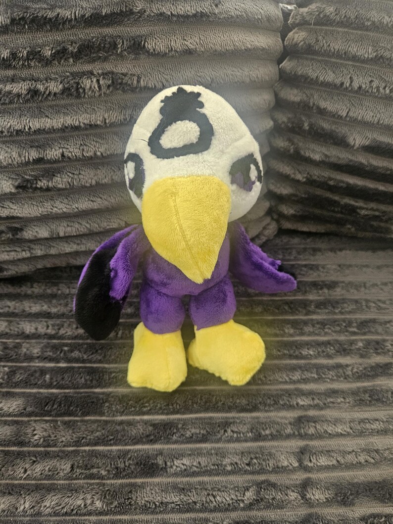 May include: A plush toy shaped like a bird with a white head, a yellow beak, and yellow feet. The body is purple with black wing tips. A black circular symbol is embroidered on the forehead. The eyes are dark with purple accents on the sides.