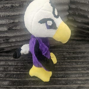May include: A plush toy shaped like a bird with a white head, a yellow beak, and purple and black wings. The toy features dark embroidered eyes and a small white tail with a black tip. The feet are yellow and the body is made of soft fabric.