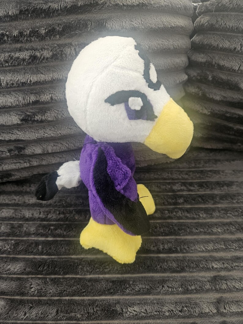 May include: A plush toy shaped like a bird with a white head, a yellow beak, and purple and black wings. The toy features dark embroidered eyes and a small white tail with a black tip. The feet are yellow and the body is made of soft fabric.