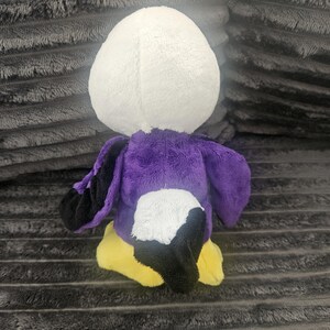 May include: A plush toy shaped like a bird with a white head, a purple body, black wing tips, a black tail, and yellow feet. The stuffed animal is constructed from soft, fuzzy fabric and is shown from a rear-facing perspective, displaying its colorful plumage.