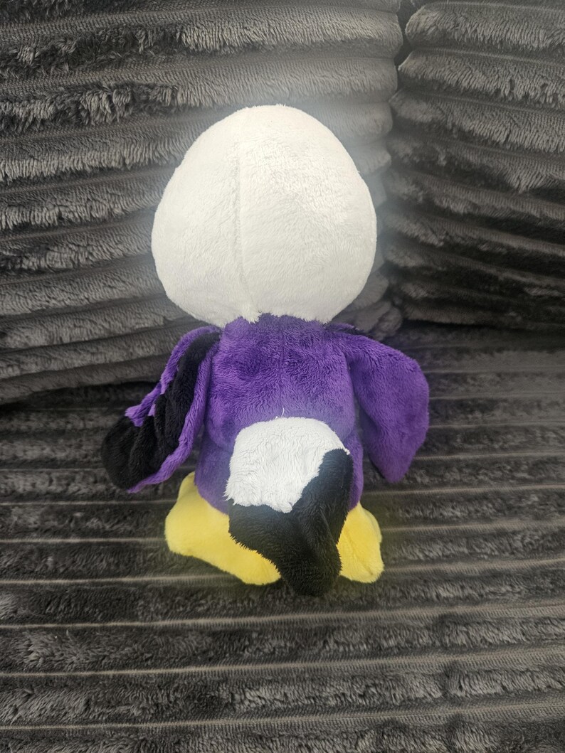 May include: A plush toy shaped like a bird with a white head, a purple body, black wing tips, a black tail, and yellow feet. The stuffed animal is constructed from soft, fuzzy fabric and is shown from a rear-facing perspective, displaying its colorful plumage.