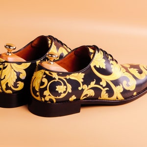May include: A pair of black leather dress shoes with a gold baroque pattern. The shoes have a classic lace-up design and are displayed with wooden shoe trees. The heels are approximately 1 inch high.