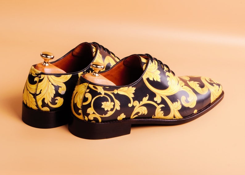 May include: A pair of black leather dress shoes with a gold baroque pattern. The shoes have a classic lace-up design and are displayed with wooden shoe trees. The heels are approximately 1 inch high.