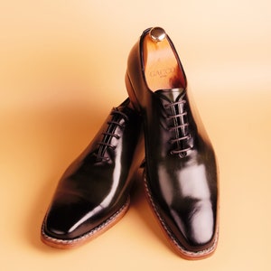 May include: A pair of black leather lace-up shoes with a polished finish. The shoes have a brown leather sole and a wooden shoe tree inside one of the shoes. The shoe tree has the word "GACCO" printed on it.