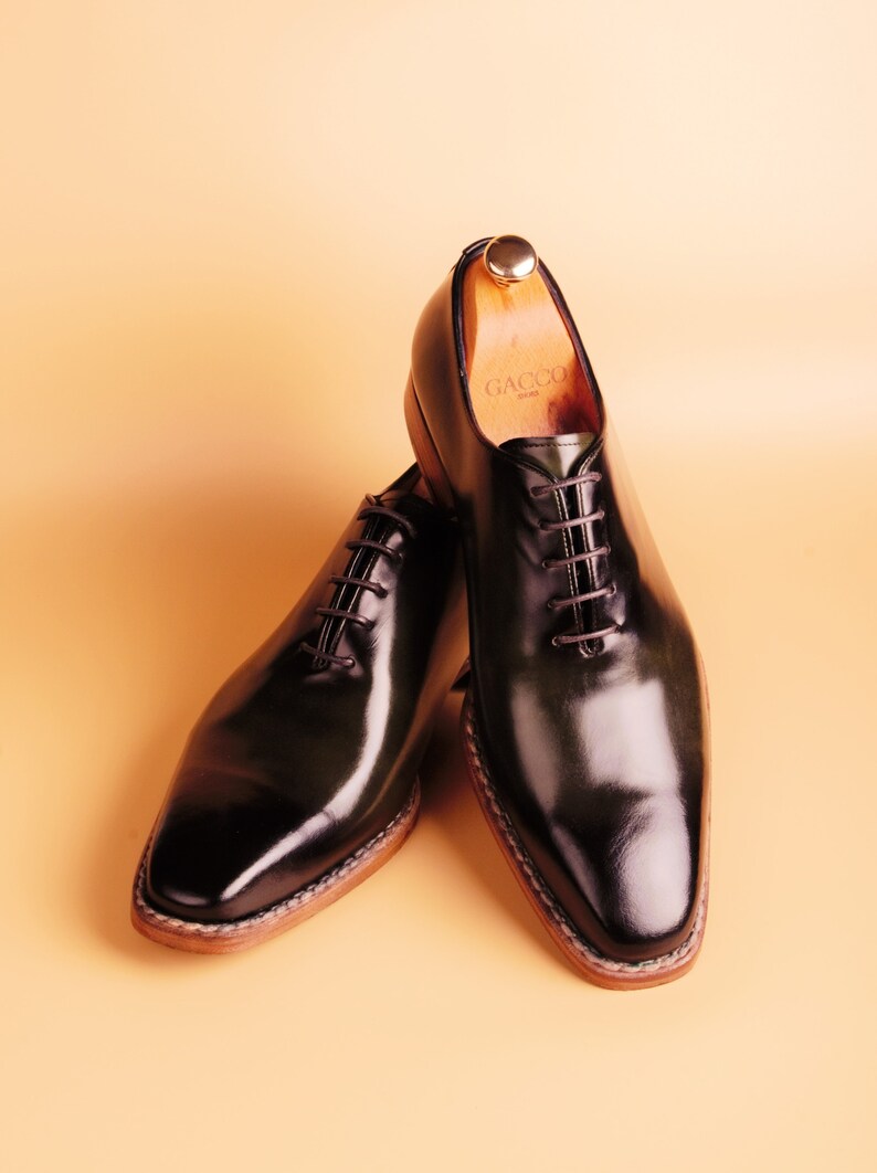 May include: A pair of black leather lace-up shoes with a polished finish. The shoes have a brown leather sole and a wooden shoe tree inside one of the shoes. The shoe tree has the word "GACCO" printed on it.