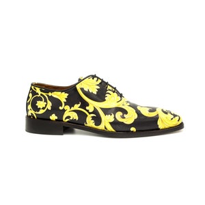 May include: Black leather dress shoe with a yellow baroque pattern. The shoe has a classic Oxford design with a lace-up closure and a low heel. The yellow pattern is a repeating floral design.