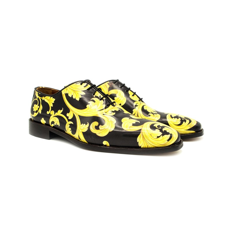 May include: A pair of black leather dress shoes with a gold baroque pattern. The shoes have black laces and a low heel. The design covers the entire shoe, including the toe and sides. The shoes are displayed on a white background.