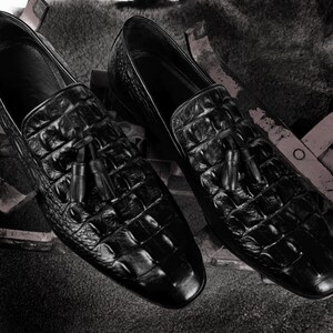 K&ouml;nnte beinhalten: Ein Paar schwarze Leder-Loafer mit Krokodilleder-Textur. Jeder Schuh hat ein Quastendetail am Blatt. Die Schuhe werden auf einer strukturierten Oberfl&auml;che pr&auml;sentiert.