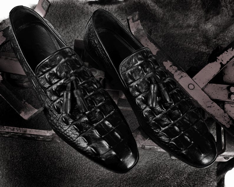 K&ouml;nnte beinhalten: Ein Paar schwarze Leder-Loafer mit Krokodilleder-Textur. Jeder Schuh hat ein Quastendetail am Blatt. Die Schuhe werden auf einer strukturierten Oberfl&auml;che pr&auml;sentiert.