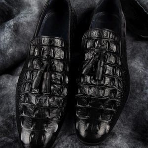 K&ouml;nnte beinhalten: Ein Paar schwarze Leder-Loafer mit Krokodilleder-Textur. Jeder Schuh hat ein Quastendetail am Blatt. Die Schuhe werden auf einer strukturierten grauen Oberfl&auml;che pr&auml;sentiert.