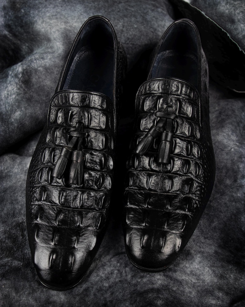 K&ouml;nnte beinhalten: Ein Paar schwarze Leder-Loafer mit Krokodilleder-Textur. Jeder Schuh hat ein Quastendetail am Blatt. Die Schuhe werden auf einer strukturierten grauen Oberfl&auml;che pr&auml;sentiert.