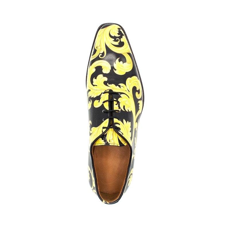May include: Black leather dress shoe with a yellow baroque pattern. The shoe has a pointed toe, black laces, and a brown interior. The design features a repeating floral scroll pattern in yellow against a black background.