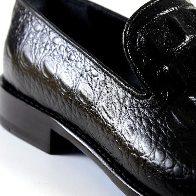 K&ouml;nnte beinhalten: Nahaufnahme eines schwarzen Lederloafers mit Krokodilhautstruktur. Der Schuh hat einen niedrigen Absatz und ein glattes, gl&auml;nzendes Finish. Das Design ist klassisch und elegant, passend f&uuml;r formelle oder gesch&auml;ftliche Kleidung.