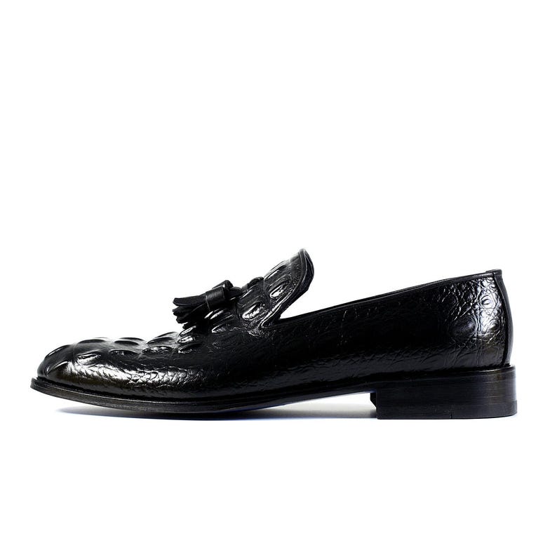 K&ouml;nnte beinhalten: Ein schwarzer Lederloafer mit einem strukturierten, gepr&auml;gten Muster. Der Schuh hat ein Quastendetail und einen niedrigen Absatz. Das Design ist elegant und klassisch, geeignet f&uuml;r formelle oder halbformelle Anl&auml;sse.