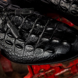 K&ouml;nnte beinhalten: Nahaufnahme eines schwarzen Lederschuhs mit Krokodillederstruktur. Der Schuh hat eine runde Spitze und ein kleines Quastendetail. Das Leder hat ein gl&auml;nzendes Finish, und der Schuh scheint neu zu sein.