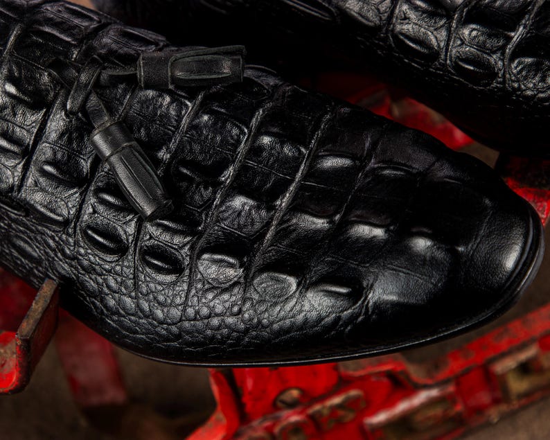 K&ouml;nnte beinhalten: Nahaufnahme eines schwarzen Lederschuhs mit Krokodillederstruktur. Der Schuh hat eine runde Spitze und ein kleines Quastendetail. Das Leder hat ein gl&auml;nzendes Finish, und der Schuh scheint neu zu sein.