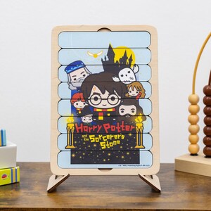 Può includere: Un puzzle in legno di Harry Potter con personaggi di "Harry Potter e la Pietra Filosofale". Il puzzle ha uno sfondo azzurro con una luna gialla e un castello nero. Il puzzle è supportato da un supporto in legno.
