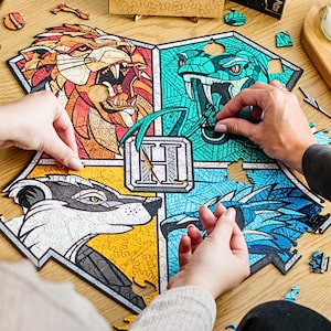 Può includere: Un puzzle a tema Harry Potter con gli stemmi delle case di Hogwarts. Il puzzle è quasi completo, mancano solo pochi pezzi. Gli stemmi sono Grifondoro, Tassorosso, Serpeverde e Corvonero. Il puzzle è su un tavolo di legno.
