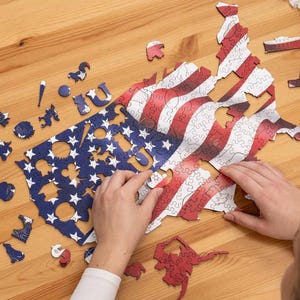 May include: A wooden jigsaw puzzle depicting the United States flag and map. The puzzle is in the process of being assembled on a wooden table. The puzzle pieces are red, white, and blue. Additional puzzle pieces are scattered around the map.